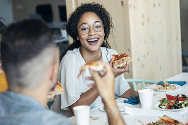 Happy woman with pizza looking at colleague in lunch break - JSRF01724