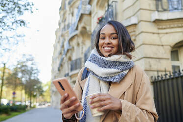 Smiling woman with mobile phone and disposable cup in city - KIJF04324