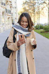 Smiling woman with coffee cup using mobile phone on footpath - KIJF04327