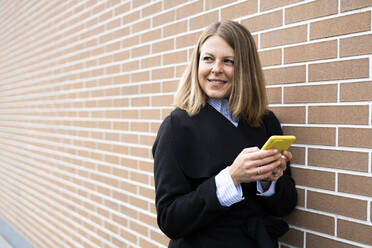 Blonde Frau hält Mobiltelefon an eine Mauer - GIOF14269