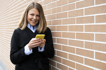 Lächelnde Frau mit Handy in der Hand und an die Wand gelehnt - GIOF14270