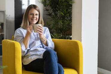 Smiling woman holding coffee cup on armchair at home - GIOF14280