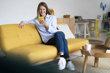 Frau hält Handy und sitzt mit gekreuzten Beinen auf dem Sofa - GIOF14283
