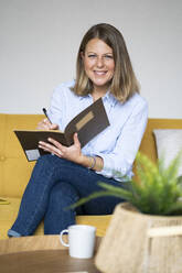 Smiling woman sitting on sofa writing in book at home - GIOF14286