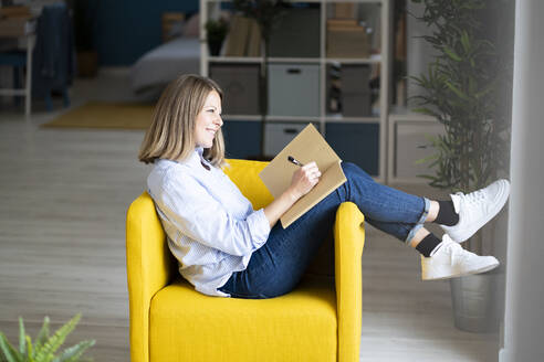 Lächelnde Frau schreibt in einem Buch auf einem Sessel zu Hause - GIOF14287