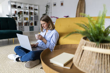 Woman with coffee cup using laptop in living room - GIOF14290