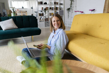 Frau mit Mobiltelefon und Laptop bei der Arbeit zu Hause - GIOF14291