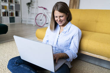 Smiling woman using laptop sitting on carpet at home - GIOF14292