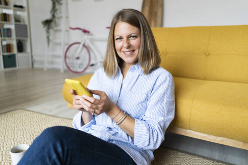 Lächelnde blonde Frau, die ein Mobiltelefon benutzt und sich zu Hause auf das Sofa lehnt - GIOF14295