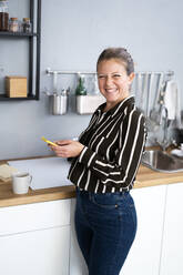 Happy woman holding mobile phone at kitchen counter - GIOF14296