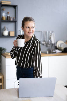 Nachdenkliche Geschäftsfrau mit Kaffeetasse und Laptop am Tisch - GIOF14297