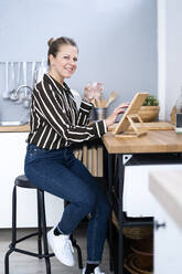 Smiling woman sitting on stool with glass of water using digital tablet in kitchen - GIOF14299