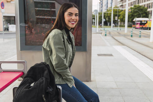 Lächelnde Frau mit Rucksack am Bahnsteig einer Straßenbahnstation - JRVF02087