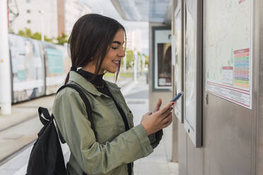 Smiling woman using mobile phone at tram station - JRVF02090