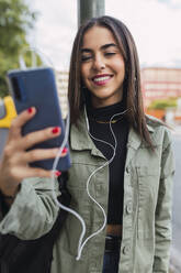 Smiling woman with in-ear headphones using mobile phone - JRVF02106