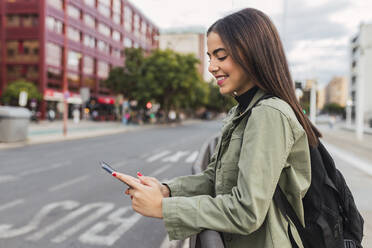 Smiling woman with backpack using mobile phone in city - JRVF02110