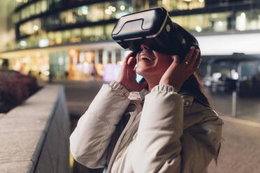 Happy young woman using virtual reality headset by wall at night - MEUF04907