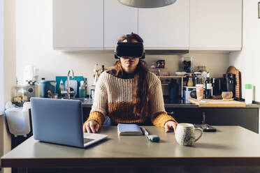 Frau mit Virtual-Reality-Headset sitzt zu Hause am Schreibtisch - MEUF04915