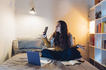 Woman doing video call though smart phone sitting with laptop on bed at home - MEUF04925