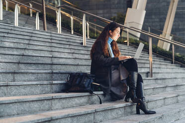 Working woman using laptop sitting on steps - MEUF04940