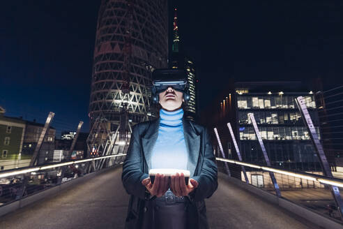 Geschäftsfrau mit Virtual-Reality-Headset hält LED-Licht auf einer Brücke in der Stadt - MEUF04965
