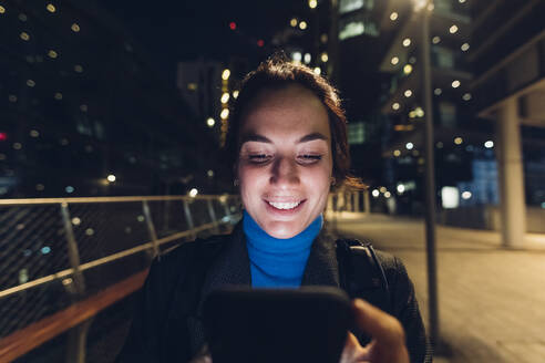 Glückliche Frau, die nachts in der Stadt ihr Smartphone benutzt - MEUF04968