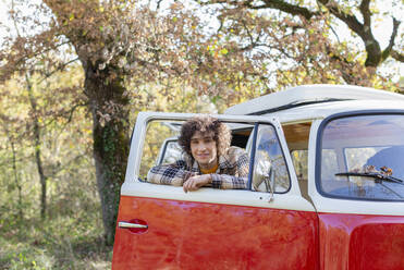 Smiling curly haired man looking through window of campervan - EIF02416