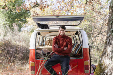Smiling young man with arms crossed sitting at campervan - EIF02435