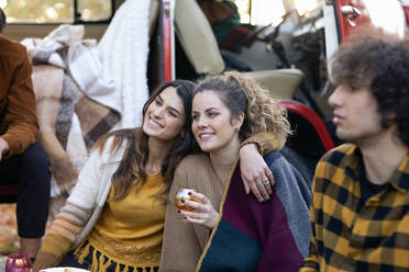 Smiling woman sitting with arm around friend on picnic outside campervan - EIF02468