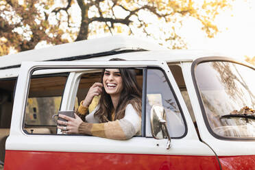 Cheerful woman with coffee mug leaning on door of van in autumn forest - EIF02480