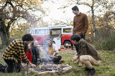 Happy friends cooking sausages in autumn forest on picnic - EIF02484