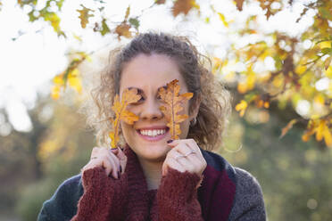 Lächelnde junge Frau hält Herbstblätter im Wald vor den Augen - EIF02487