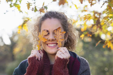 Smiling young woman holding autumn leaves in front of eyes at forest - EIF02487