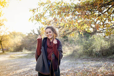 Smiling beautiful woman holding autumn leaves in forest on weekend - EIF02488