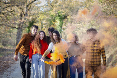 Cheerful woman enjoying smoke flare with friends in autumn forest - EIF02496