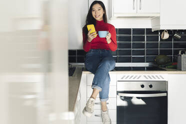 Smiling woman with coffee cup and mobile phone at kitchen counter - JCCMF04729
