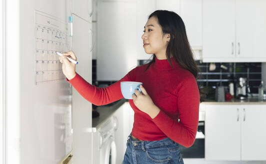 Young woman writing on calendar in kitchen - JCCMF04730