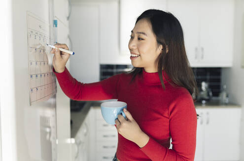 Smiling woman with coffee cup writing on calendar in kitchen - JCCMF04731