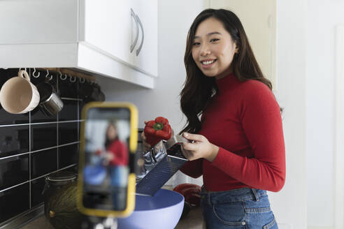Influencer with grate and vegetable filming through mobile phone in kitchen - JCCMF04733