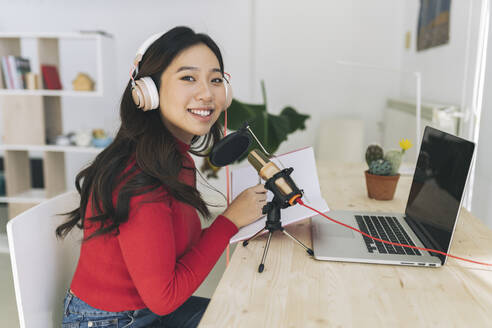 Lächelnde Frau mit Kopfhörern und Laptop beim Podcasten zu Hause - JCCMF04737