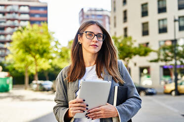 Seriöse Arbeitnehmerin mit Brille surft auf einem Tablet, während sie auf der Straße steht und ihre Arbeit erledigt - ADSF32124