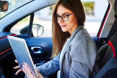 Side view of crop smiling well dressed female worker in eyeglasses sitting in car and watching tablet - ADSF32125