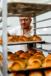 Männlicher Bäcker mit Tattoos auf dem Arm backt Croissants in einem großen Metallofen während der Arbeit in einer Bäckerei - ADSF32131