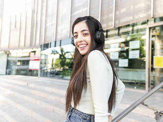 Happy woman with long brown hair in casual clothes adjusting headphones and looking at camera on street against modern building in city in daylight - ADSF32135