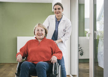 Smiling nurse standing behind woman in wheelchair at home - UUF25104