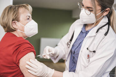 Ein Arzt mit Gesichtsmaske injiziert einem Patienten zu Hause eine Impfung - UUF25109