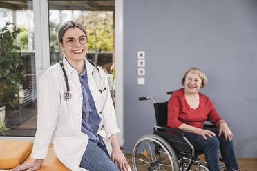 Happy doctor sitting with woman in wheelchair at home - UUF25120