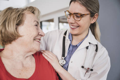 Smiling doctor with hand on senior patient's shoulder at home - UUF25127