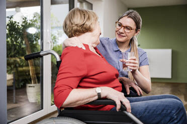 Smiling care assistant giving water to disabled woman at home - UUF25139