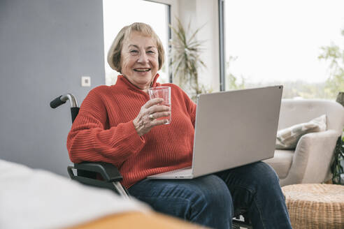 Lächelnde behinderte Frau mit Trinkglas und Laptop im Rollstuhl - UUF25198